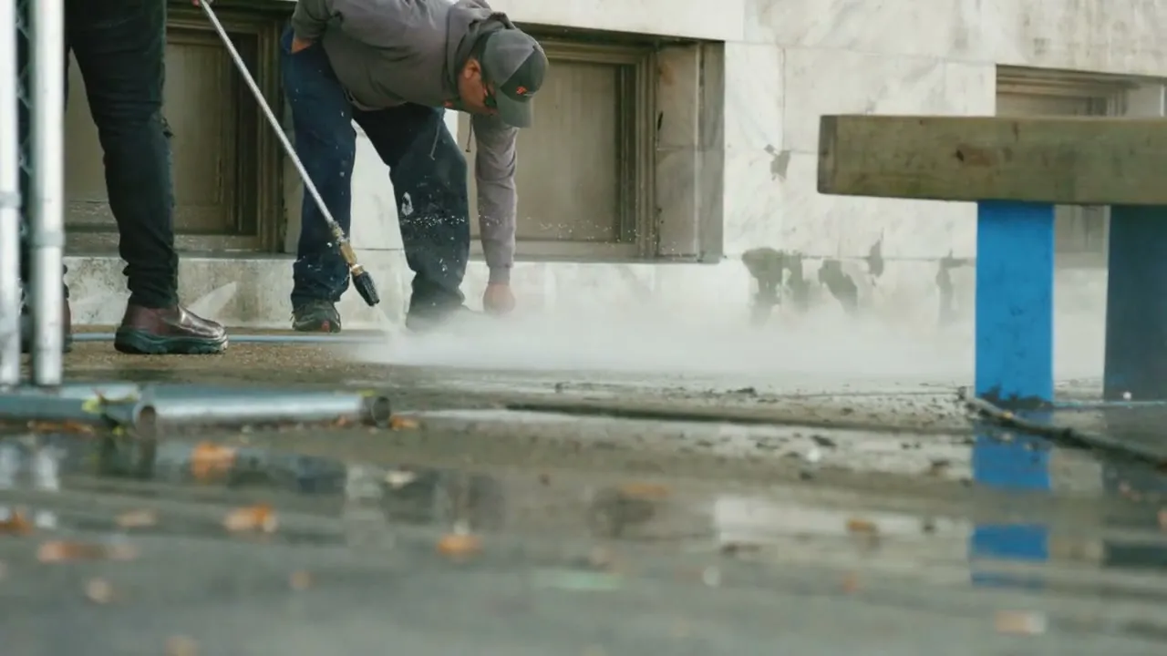 Medewerker reinigt een betonnen buitenvloer met de hogedrukreiniger, met zichtbare waternevel