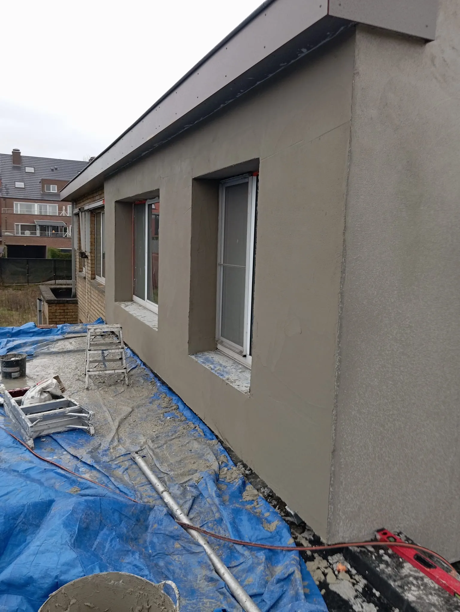Uitbouw met grijze basispleisterlaag over de isolatie, met werfzeil op het platte dak