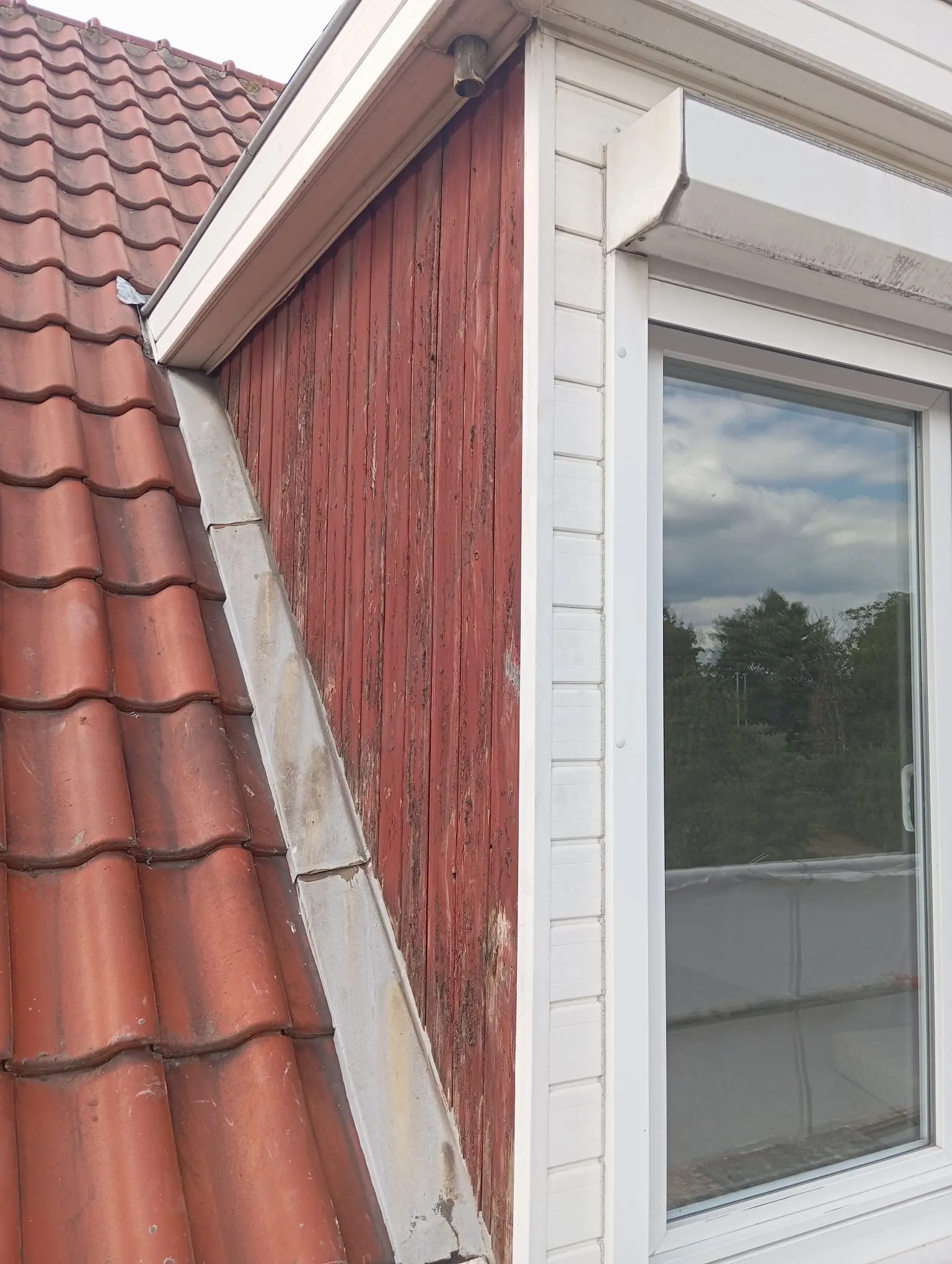 Dakkapel met verweerde rode houten beplanking naast een pannendak, voor de renovatie